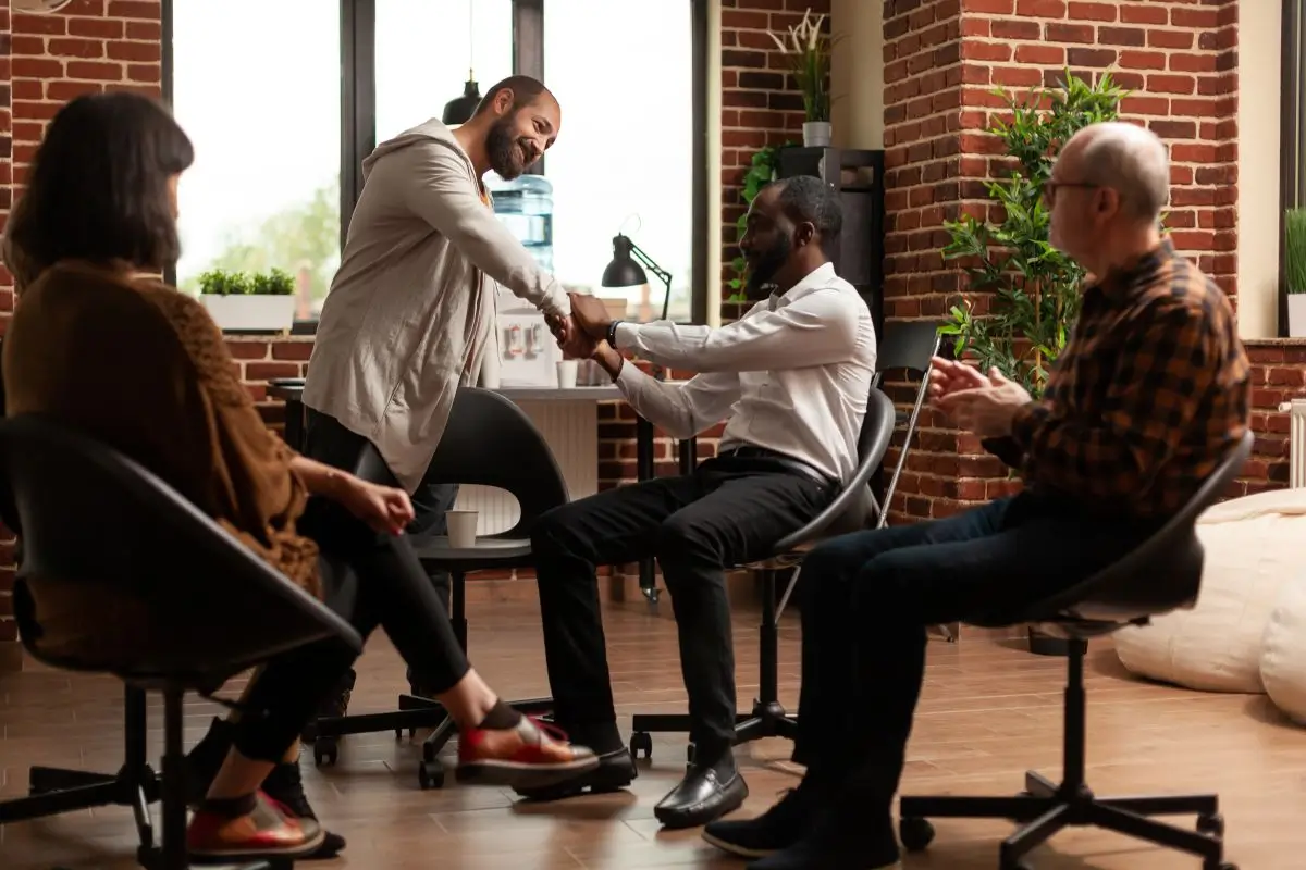 Support group meeting with diverse individuals sitting in a circle, shaking hands and showing encouragement during a recovery or counseling session.
