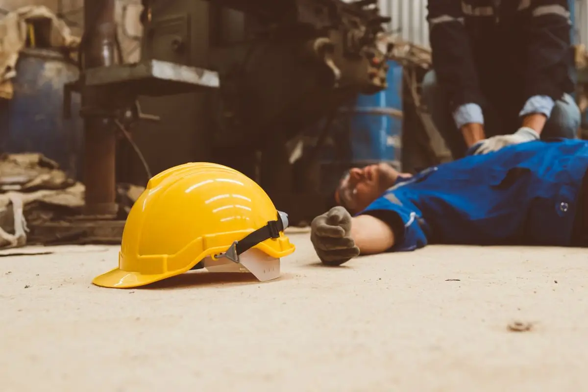 Injured worker lying on the factory floor after an industrial accident with a yellow safety helmet nearby.