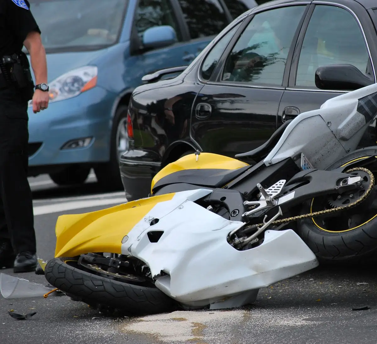 Damaged yellow and white motorcycle lying on the road after a collision with a car, with a police officer at the scene.