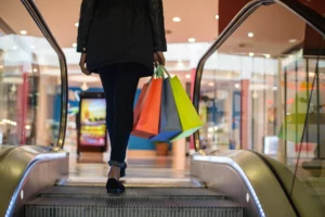 Person in a black coat and jeans riding an escalator in a mall with colorful shopping bags — capturing a vibrant shopping scene and underscoring the premises-liability risks discussed in Massachusetts mall and retail settings.