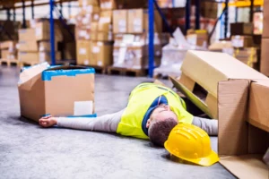 Stressed warehouse worker lying on the floor among fallen boxes with a yellow hard hat nearby — conveying a workplace accident, potential injury, and the risks of warehouse hazards that may lead to a workers’ compensation or personal-injury claim.